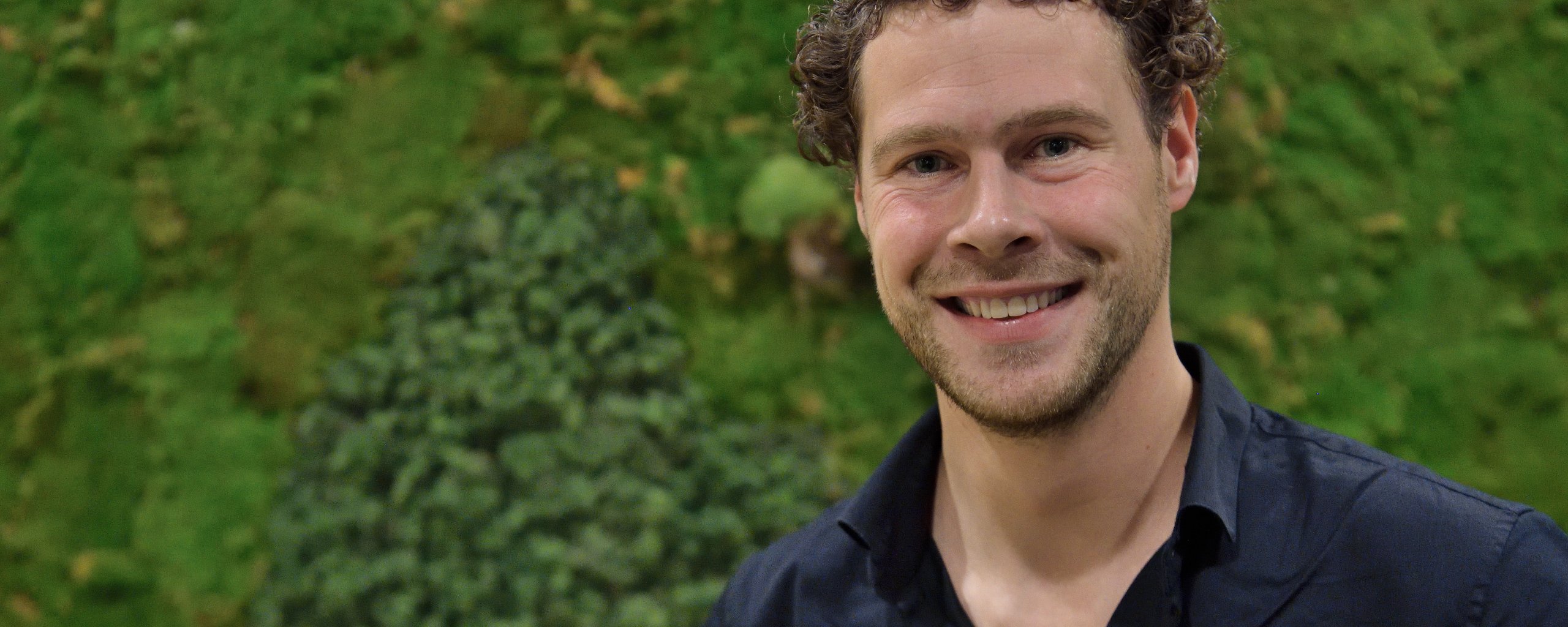 Een lachende man staat voor een wand bekleed met mos.